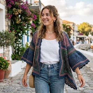 May include: A woman wearing a blue, green, and pink crochet cardigan over a white top and blue jeans. She is walking on a cobblestone street with buildings and flowers in the background. A straw bag hangs from her shoulder.
