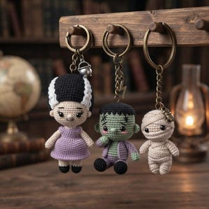 May include: Three crochet keychains featuring a bride figure with black and white hair in a purple dress, a green monster figure with a lightning bolt charm, and a beige mummy figure, all attached to bronze-toned metal rings and chains.