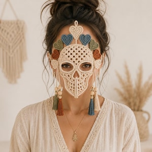 May include: A macrame skull mask crafted from cream-colored cord, featuring a detailed woven pattern. The top is adorned with a row of small, multi-colored hearts in blue, green, and rust. Two long, dangling tassels with wooden beads hang down.