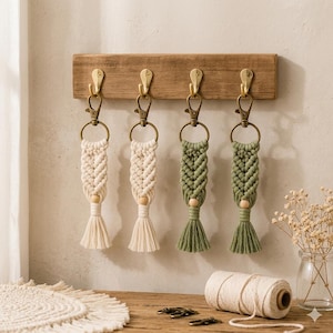 May include: Four macrame keychains hanging from a wooden wall rack with brass hooks. Two keychains are cream-colored and two are sage green. Each features a braided cord pattern, a single wooden bead, a tassel, and a brass lobster claw clasp.