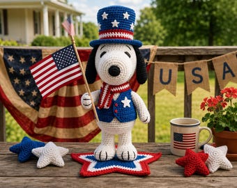 Op de afbeelding: Een gehaakt wit hondenfiguur met een blauwe hoge hoed met witte sterren, een blauw vest met een witte ster en een rood-wit gestreepte sjaal. Het figuur houdt een kleine Amerikaanse vlag vast en staat op een stervormige mat in rood, wit en blauw.