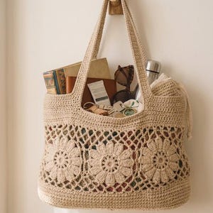 Könnte beinhalten: Beige gehäkelte Tragetasche mit floralem Muster, an einem Holzhaken hängend. Die Tasche enthält Bücher, Sonnenbrillen und eine Wasserflasche. Die Tasche ist ca. 38 cm breit und 30 cm hoch.