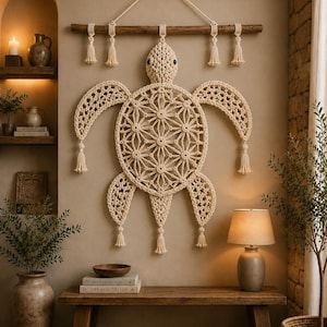Op de afbeelding: Een grote off-white macramé wandhanger in de vorm van een zeeschildpad, opgehangen aan een horizontale houten tak. De schildpad heeft een ingewikkeld geometrisch patroon op het schild, donkere ogen en decoratieve kwastjes aan de vinnen.