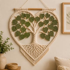 May include: A heart-shaped macrame wall hanging featuring a central tree design with off-white twisted cotton cord branches and roots. The tree is adorned with numerous small, textured sage green crocheted leaves and light wooden beads.