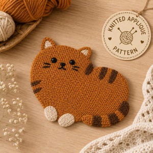 Puede incluir: Una aplicación de gato naranja tejido con detalles faciales negros y rayas marrones. El gato tiene patas y orejas blancas. Una etiqueta de madera con el texto "KNITTED APPLIQUE PATTERN" cuelga cerca. Hilo y un artículo de punto en el fondo.