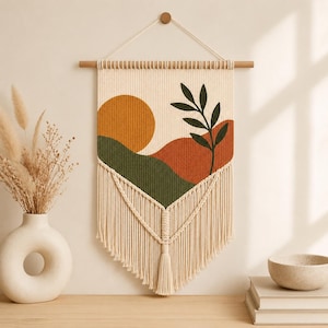 May include: A cream-coloured macrame wall hanging suspended from a wooden dowel. The textile features a printed landscape design with a yellow sun, green hills, and a rust-coloured slope, accented by a dark green leafy branch and long fringe.