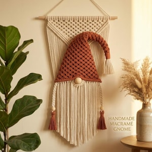 May include: A wall hanging featuring a macramé gnome with a rust-colored textured hat and a long cream-colored fringe beard. The gnome is suspended from a wooden dowel by a cream macramé cord, with two braided side cords ending in tassels.