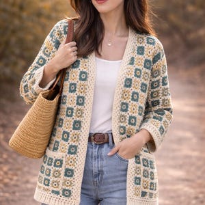 May include: A cream-coloured, open-front crochet cardigan with a geometric pattern in teal and yellow. The cardigan is paired with a white top, blue jeans, and a brown belt. A woven tote bag is carried over the shoulder.