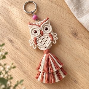 Puede incluir: Un llavero de búho en macramé con cordón color crema y terracota, dos ojos de cuentas marrón oscuro y una cuenta rosa sobre el búho. El diseño incluye una borla en capas en la parte inferior y una anilla de metal plateado.