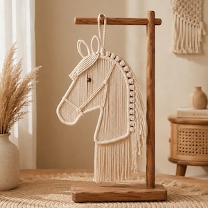 May include: A cream-colored macrame horse head wall hanging suspended from a wooden stand. The horse features a braided rope mane, a single dark brown bead for an eye, and a halter design crafted from knotted cotton cord with a loop at the top.