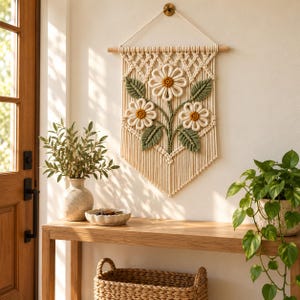 Può includere: Un arazzo in macramè color crema con tre fiori bianchi simili a margherite, centri giallo senape e foglie verde salvia. Il design è appeso a un bastone di legno orizzontale e presenta un motivo a nodi a forma di rombo.