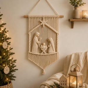 Puede incluir: Un tapiz de macramé color crema que representa una escena de natividad. El diseño muestra dos figuras de pie y arrodilladas junto a una cuna central, enmarcadas por una estructura triangular rematada con una estrella y flecos en la base.