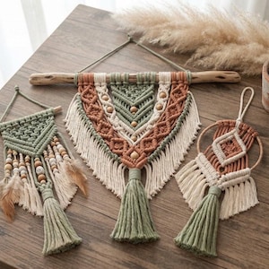 Puede incluir: Tres tapices de macramé hechos con cuerda de algodón en color verde salvia, terracota y crema. Los diseños incluyen patrones de nudos intrincados, cuentas de madera, flecos y borlas. Una pieza cuelga de una rama de madera y otra de un aro.