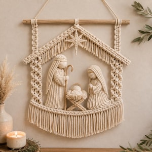 May include: A cream-colored macrame wall hanging featuring a nativity scene with three figures and a star. The structure is suspended from a wooden dowel and includes a central manger, a standing figure with a staff, and a kneeling figure.