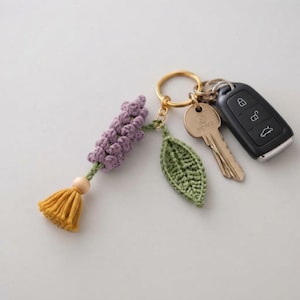 May include: A gold-toned metal keyring featuring a crocheted purple lavender sprig, a green crocheted leaf, and a mustard yellow tassel with a wooden bead. Attached to the ring are a brass-coloured metal key and a black plastic car key fob.