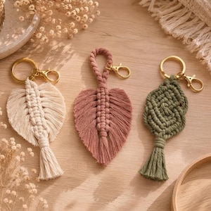 Può includere: Tre portachiavi in macramè con moschettoni e anelli in metallo dorato. Due portachiavi hanno la forma di una foglia nei colori crema e rosa antico, mentre il terzo presenta un design annodato verde. Ogni pezzo include una frangia centrale.