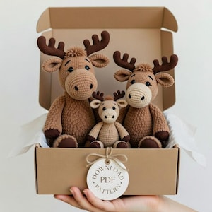 Puede incluir: Tres juguetes de alce de ganchillo en una caja de cartón. Los alces son marrones con caras color crema y cornamentas marrones oscuras. Un pequeño alce se sienta entre dos más grandes. Una etiqueta con "DOWNLOAD PDF PATTERN" está adjunta.
