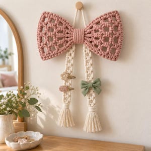 May include: A macrame wall hanging shaped like a large dusty pink bow with two long cream-colored vertical strips. The strips feature attached hair clips, including a flower, a textured oval, and a small sage green fabric bow.