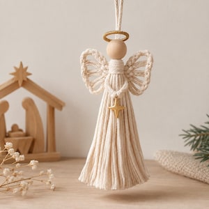 May include: A macrame angel ornament crafted from off-white cotton cord, featuring a smooth wooden bead head, a gold-toned wire halo, and a gold-toned four-pointed star charm suspended at the center of the body with fringed skirt detailing.