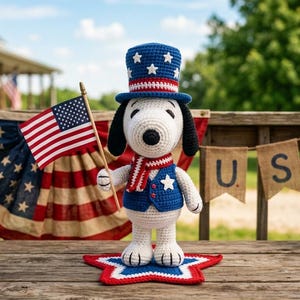 May include: A crocheted Snoopy figure dressed in patriotic attire, holding a miniature American flag. He wears a blue vest with a white star, a red, white, and blue scarf, and a matching top hat. The scene is decorated with USA banners and star-shaped accents.