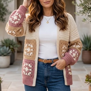 May include: A cream, tan, and pink crochet cardigan with a square pattern design. The open-front sweater has long sleeves and a brown belt. The sleeves and front panels feature a floral-like pattern. The cardigan is worn over a white top and blue jeans.