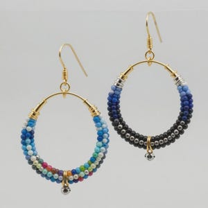 May include: Two pairs of hoop earrings. One pair features a gradient of blue, white, and colorful beads. The other pair has a gradient of black and blue beads. Both have gold-colored metal accents and a small dangling charm.