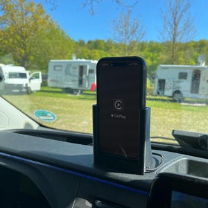May include: A black smartphone displaying the CarPlay interface is mounted in a car. The phone is in a black holder on the dashboard. The background shows a campsite with caravans and trees under a blue sky.