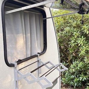 Peut inclure: Un étendoir à linge pliable blanc et gris monté sur le côté d'une caravane. Le séchoir est déployé, prêt à l'emploi. Une fenêtre avec un rideau est visible derrière le séchoir.