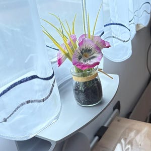 Könnte beinhalten: Ein kleines Glasgefäß, gefüllt mit dunklen Kieselsteinen, mit rosa und weißen Kunstblumen mit gelben Stielen. Das Glas ist mit einem dünnen, hellbraunen Band gebunden. Die Anordnung steht auf einem weißen Regal in der Nähe eines Fensters mit transparenten Vorhängen.