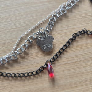 May include: A layered necklace featuring a silver-toned chain and a black chain. The silver chain includes a Mickey Mouse-shaped charm with the text Disney and China engraved on it. The black chain features a single faceted red teardrop pendant.