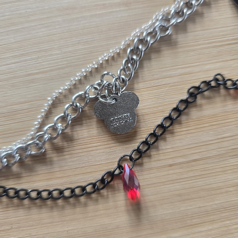 May include: A layered necklace featuring a silver-toned chain and a black chain. The silver chain includes a Mickey Mouse-shaped charm with the text Disney and China engraved on it. The black chain features a single faceted red teardrop pendant.