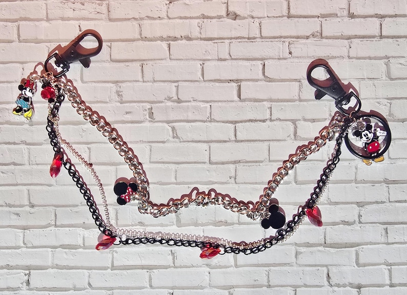 May include: A multi-strand decorative chain featuring two black lobster clasps at the ends. The design includes silver and black metal links, red crystal accents, and small charms depicting cartoon mouse characters in red, black, and yellow colors.