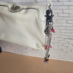 May include: A white leather handbag featuring a silver-tone metal turn-lock closure and a white strap. Attached to the strap is a decorative keychain composed of silver and black chains, black plastic clips, and small red translucent beads.