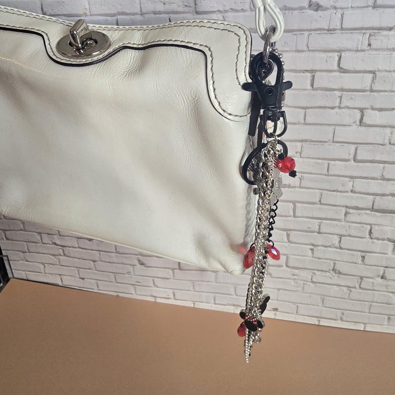 May include: A white leather handbag featuring a silver-tone metal turn-lock closure and a white strap. Attached to the strap is a decorative keychain composed of silver and black chains, black plastic clips, and small red translucent beads.
