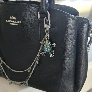 Puede incluir: Bolso Coach de cuero negro con un charm de rana plateada. El charm está adornado con pedrería azul y verde y cuelga de una cadena plateada. El bolso tiene un asa superior y un logotipo plateado de Coach.