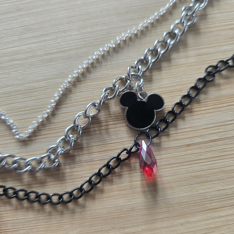 May include: A layered necklace featuring a delicate silver chain, a thicker silver curb chain with a black mouse-ear silhouette charm, and a thin black chain adorned with a single faceted, translucent red teardrop-shaped crystal pendant.