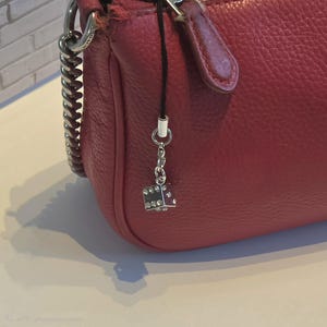 May include: A textured red leather handbag featuring a silver chain strap and a dangling charm accessory. The charm consists of a black cord attached to a small, silver-toned cube shaped like a die, which is embellished with clear sparkling crystals.