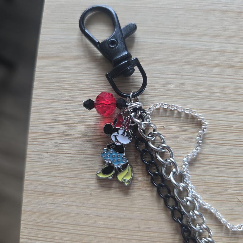 May include: A keychain featuring a black lobster clasp, a Minnie Mouse charm with a red bow, blue polka-dot skirt, and yellow shoes. The accessory includes a red faceted bead, a black bead, a black metal chain, and a silver-toned link chain.
