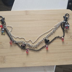 May include: A multi-layered chain accessory featuring silver and black metal links, two black lobster clasps, and several charms including Mickey and Minnie Mouse figures, black mouse ear silhouettes, and small translucent red teardrop beads.
