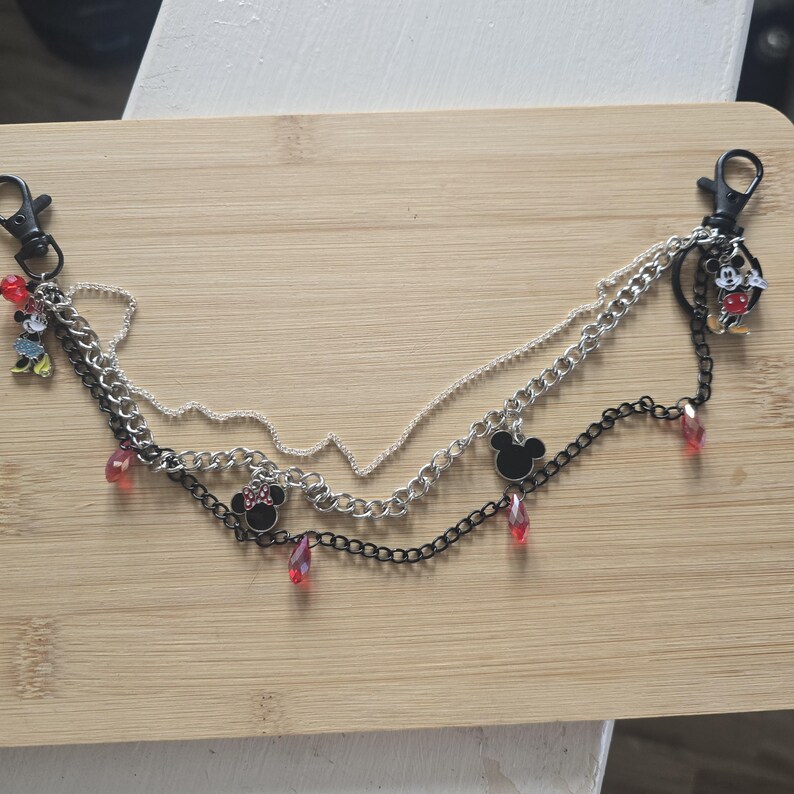 May include: A multi-layered chain accessory featuring silver and black metal links, two black lobster clasps, and several charms including Mickey and Minnie Mouse figures, black mouse ear silhouettes, and small translucent red teardrop beads.
