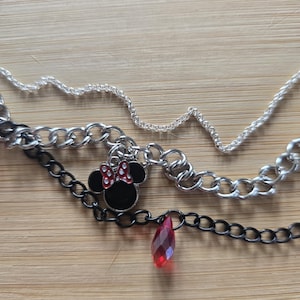 May include: A multi-layered necklace featuring a thin silver chain, a thicker silver curb chain, and a black chain. The necklace includes a black and red enamel mouse-shaped charm with a bow and a single faceted red teardrop-shaped pendant.
