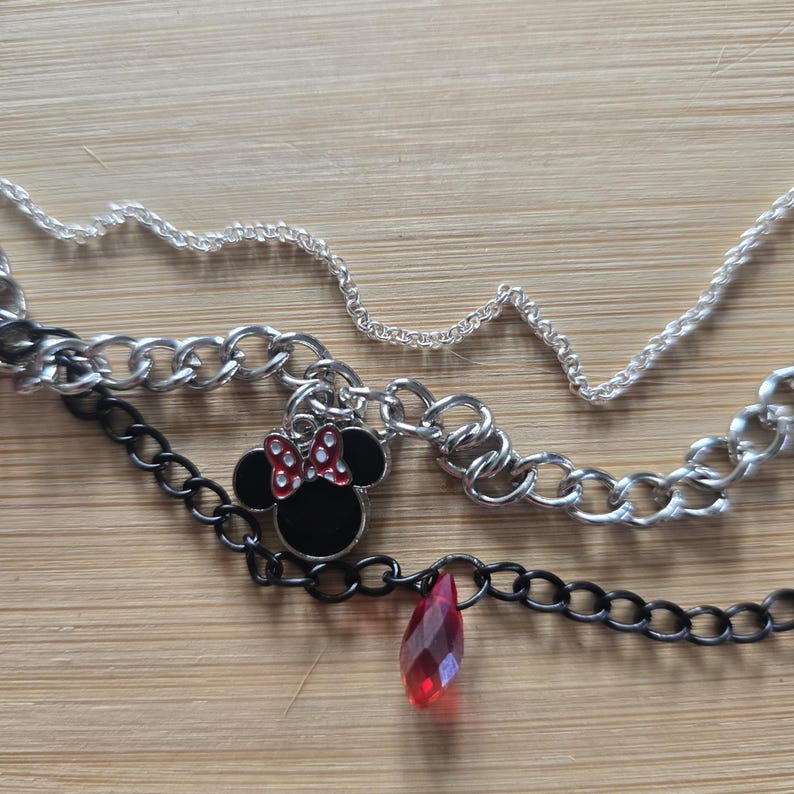 May include: A multi-layered necklace featuring a thin silver chain, a thicker silver curb chain, and a black chain. The necklace includes a black and red enamel mouse-shaped charm with a bow and a single faceted red teardrop-shaped pendant.