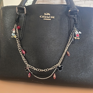 May include: A black textured leather Coach handbag adorned with a decorative double-layered silver-tone chain accessory. The chain features dangling charms of Mickey and Minnie Mouse, black enamel mouse ears, and faceted red crystal beads.