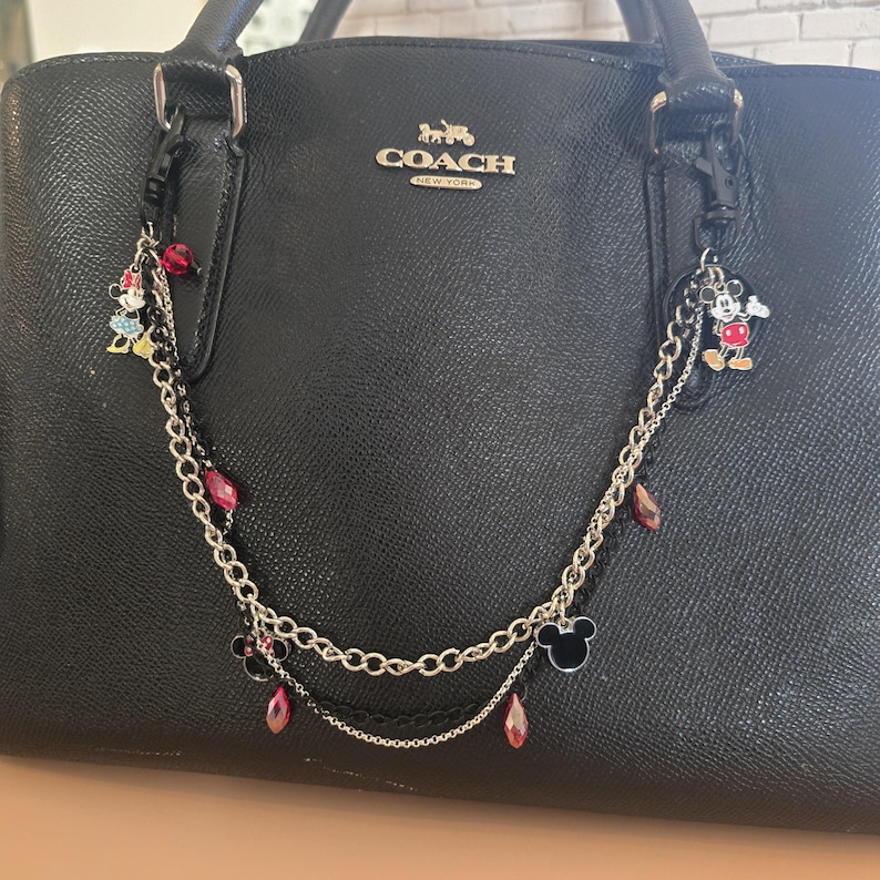 May include: A black textured leather Coach handbag adorned with a decorative double-layered silver-tone chain accessory. The chain features dangling charms of Mickey and Minnie Mouse, black enamel mouse ears, and faceted red crystal beads.
