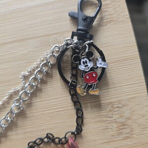 May include: A keychain featuring a black lobster clasp attached to a black circular ring. A silver-tone chain and a black chain hang from the ring. A small enamel charm of Mickey Mouse in black, white, red, and yellow is attached to the black ring.