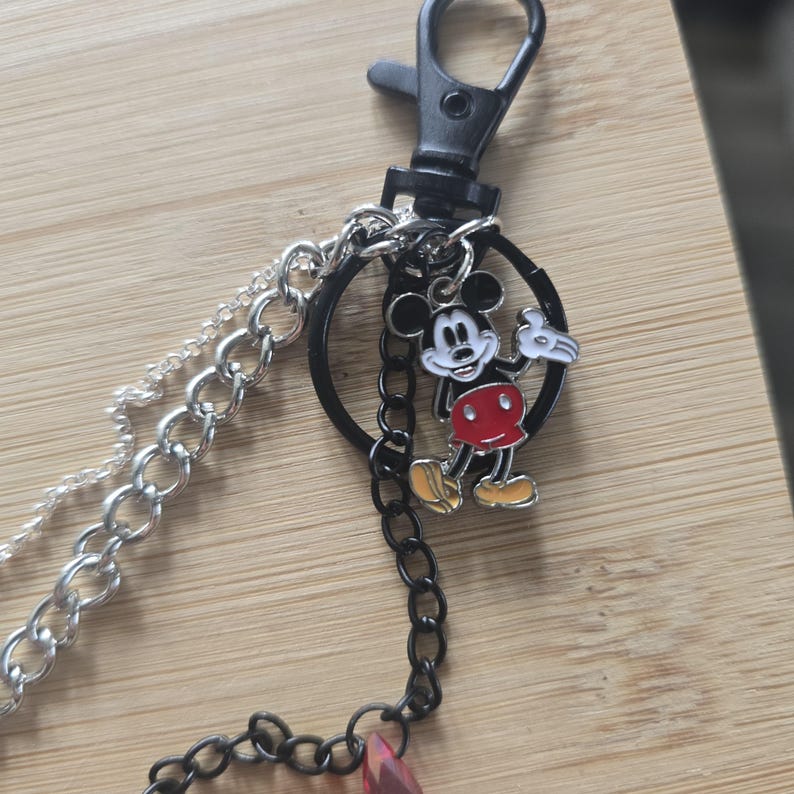May include: A keychain featuring a black lobster clasp attached to a black circular ring. A silver-tone chain and a black chain hang from the ring. A small enamel charm of Mickey Mouse in black, white, red, and yellow is attached to the black ring.