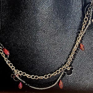 May include: A decorative gold-tone metal chain accessory for a handbag, featuring dangling charms of Mickey Mouse and Minnie Mouse, interspersed with small red faceted beads and a black Mickey Mouse silhouette icon against a black textured bag.