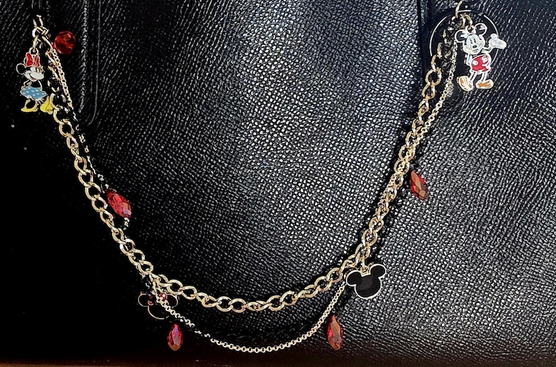 May include: A decorative gold-tone metal chain accessory for a handbag, featuring dangling charms of Mickey Mouse and Minnie Mouse, interspersed with small red faceted beads and a black Mickey Mouse silhouette icon against a black textured bag.