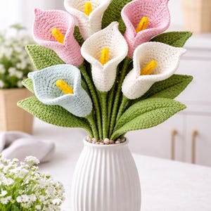 May include: A handcrafted bouquet of crochet calla lilies in a white vase. The flowers are in shades of pink, white, and blue, with yellow centers and green leaves. The vase is white with vertical ridges, creating a textured appearance.