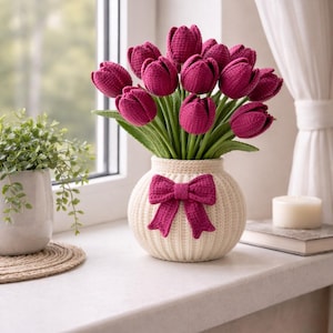Può includere: Un bouquet di tulipani magenta all'uncinetto in un vaso color crema con un fiocco magenta. Il vaso è su un davanzale, accanto a una piccola pianta in vaso e una candela. Sullo sfondo, una finestra e una tenda bianca.